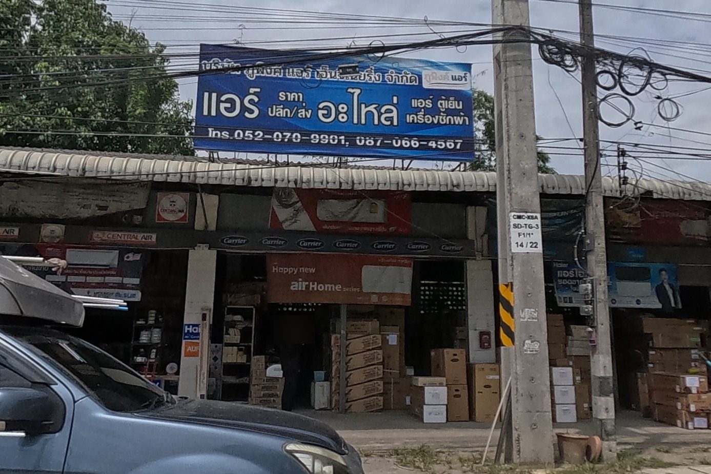 A wholesale air-conditioner, fridge and washing-machine parts shop with a blue sign and phone numbers, boxes of stock stacked outside, a customized pickup truck parked in front.