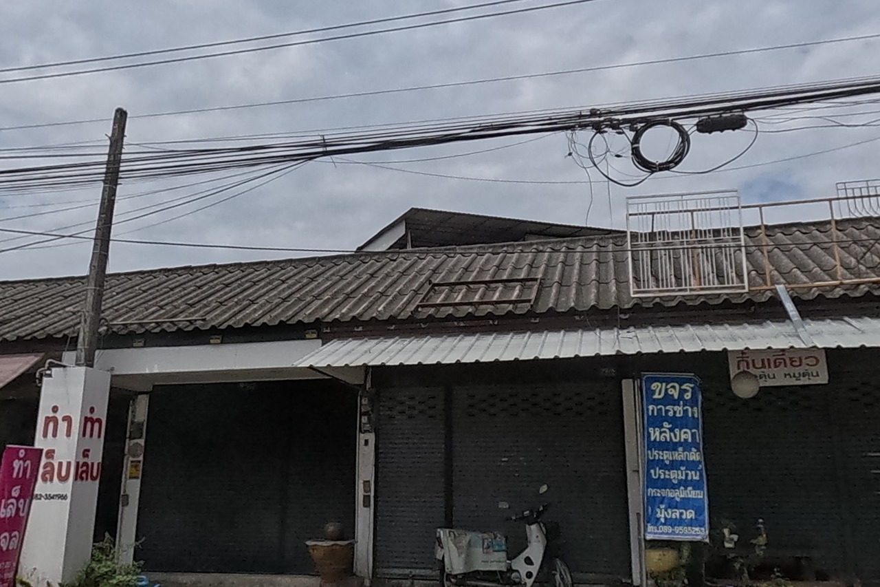 A row of shophouses: a metalwork/repair shop with a blue sign and phone number, a gel-nail shop's pink sign, and a small restaurant with potted plants and a wooden carving out front.