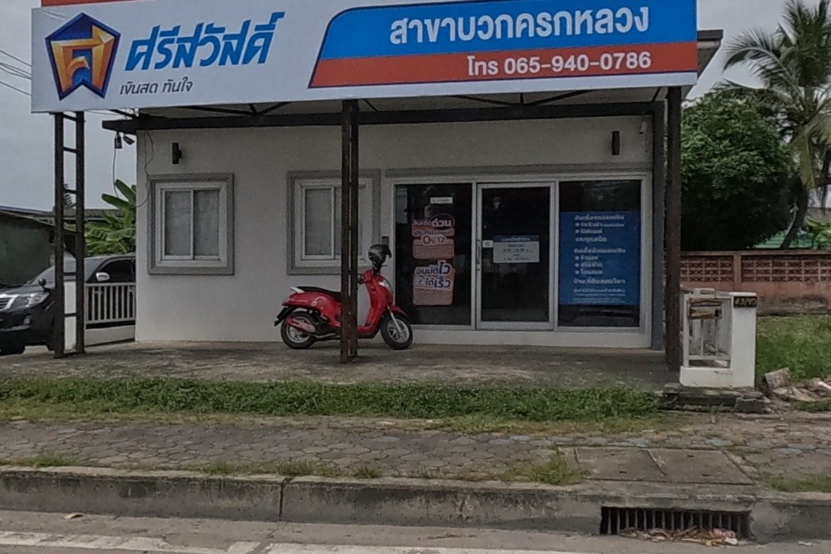A Srisawad branch office under a large red billboard for car-insurance and loan instalments, a small shrine and potted flowers by the pole, a red scooter parked out front.