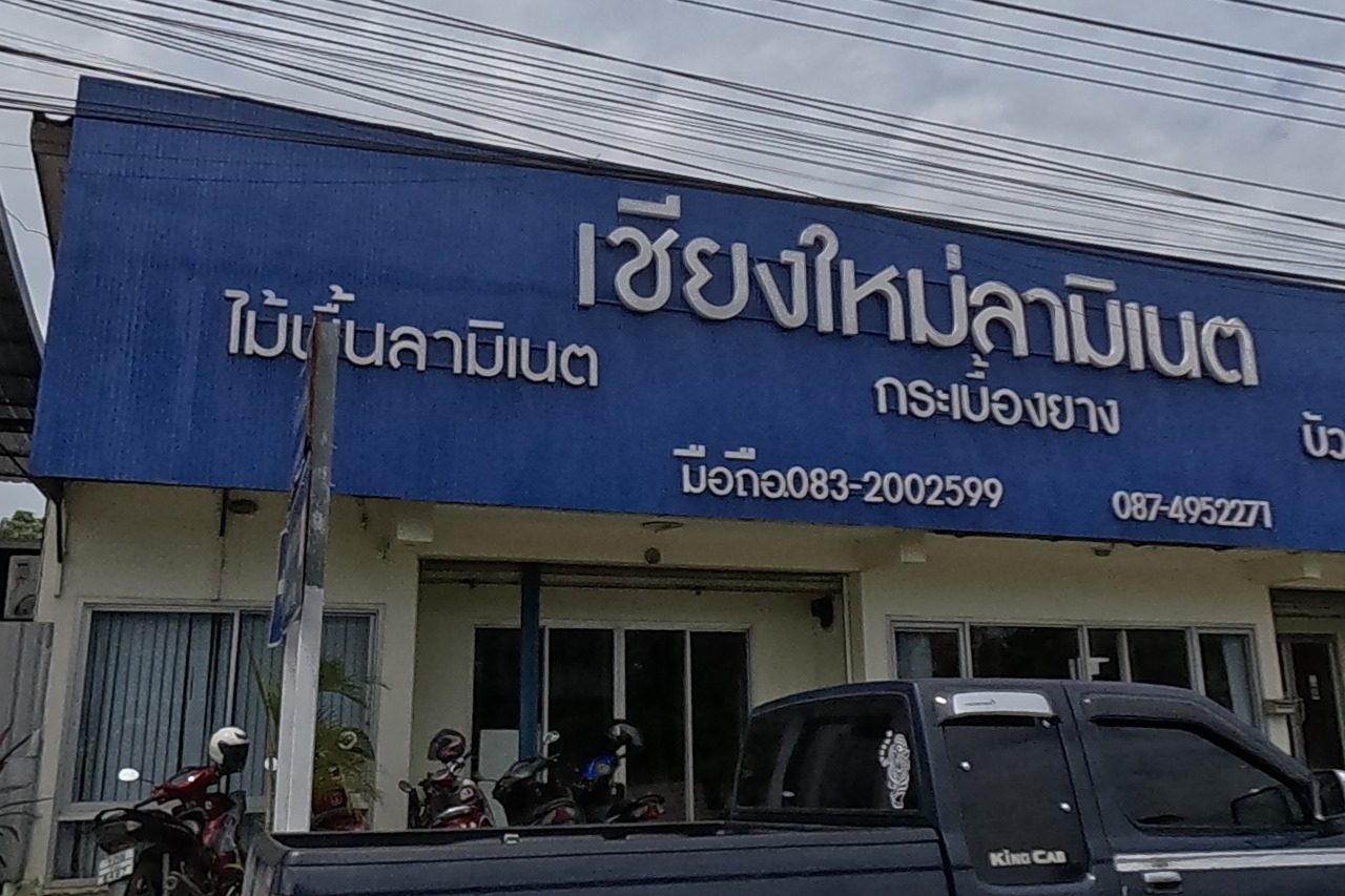 A large blue-walled warehouse for a laminate flooring and vinyl tile shop, sign with two phone numbers, staff standing at the roller-shutter entrance, a dark pickup parked out front.