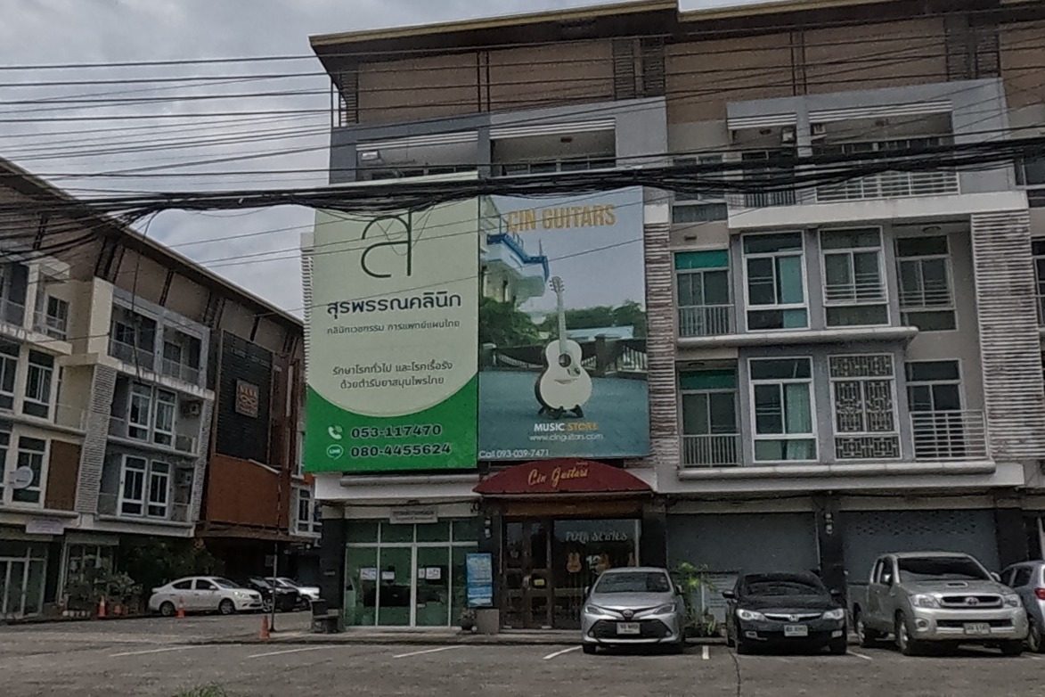 A row of modern shophouses: a clinic's green banner, a guitar shop with a large guitar-store banner, a property developer's red-signed office, and more shops beyond, cars parked in front, a train-station directional sign by the road.