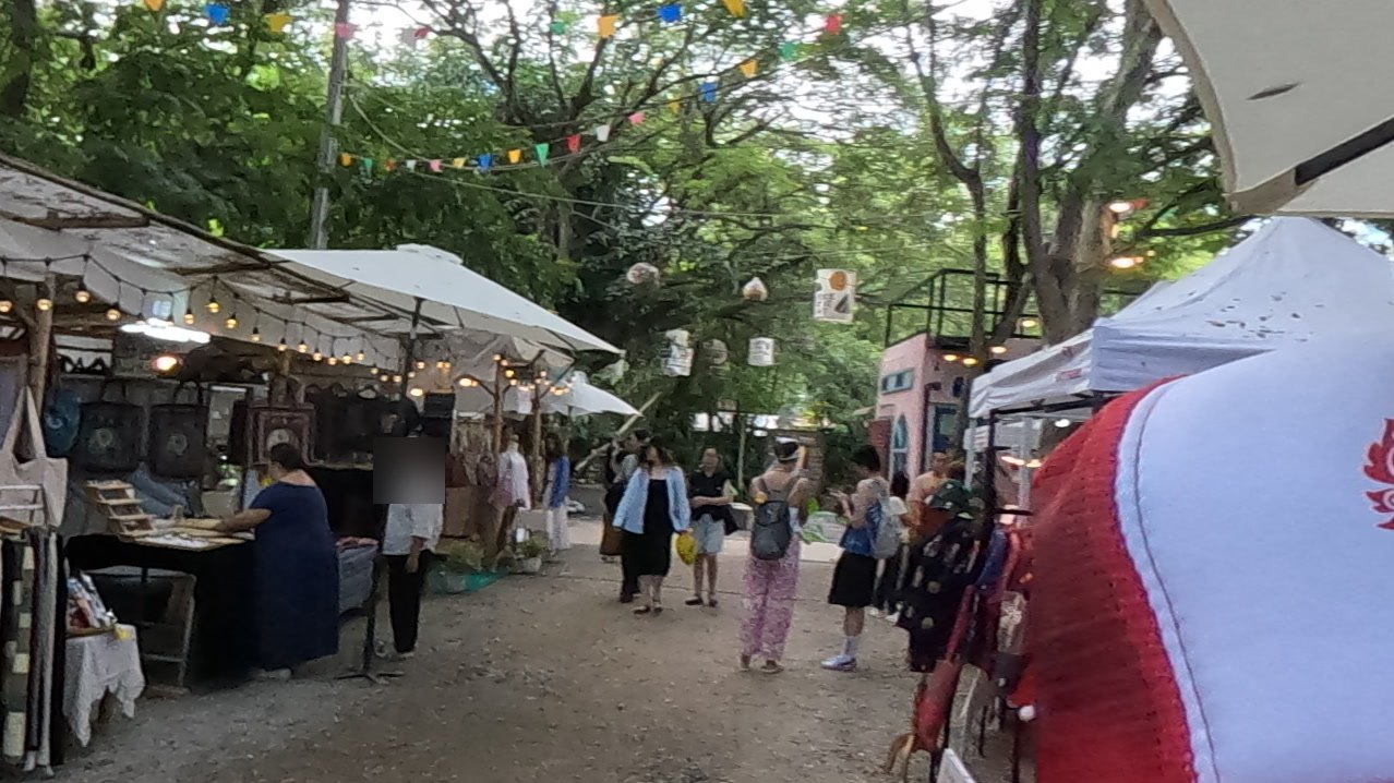 The stall lane under rain trees and bunting — Chamcha Market, San Kamphaeng