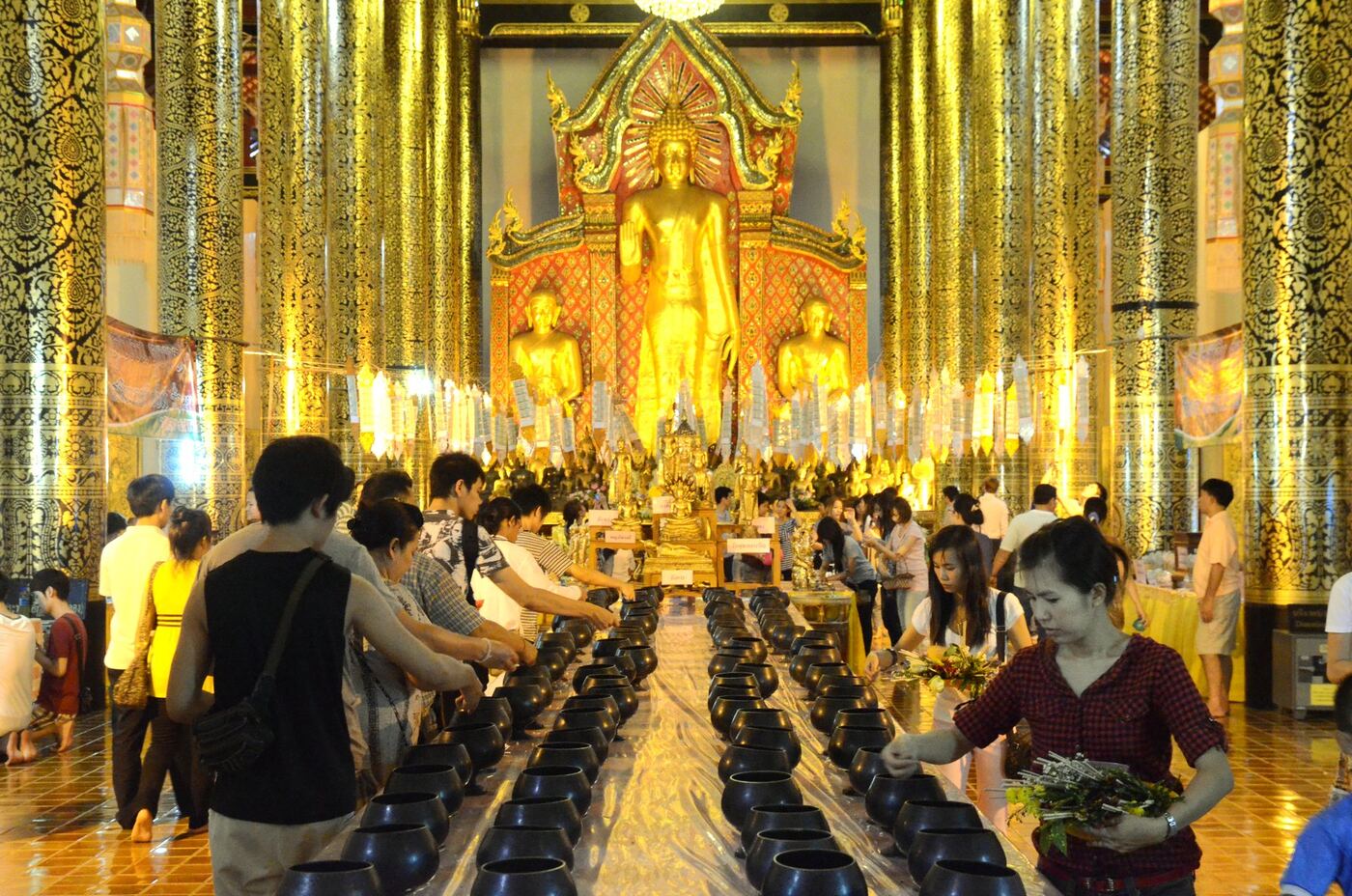 People move between two long rows of black alms bowls on the floor of a gold-pillared hall and drop coins into them in front of a standing Buddha; one woman holds a bunch of flowers.