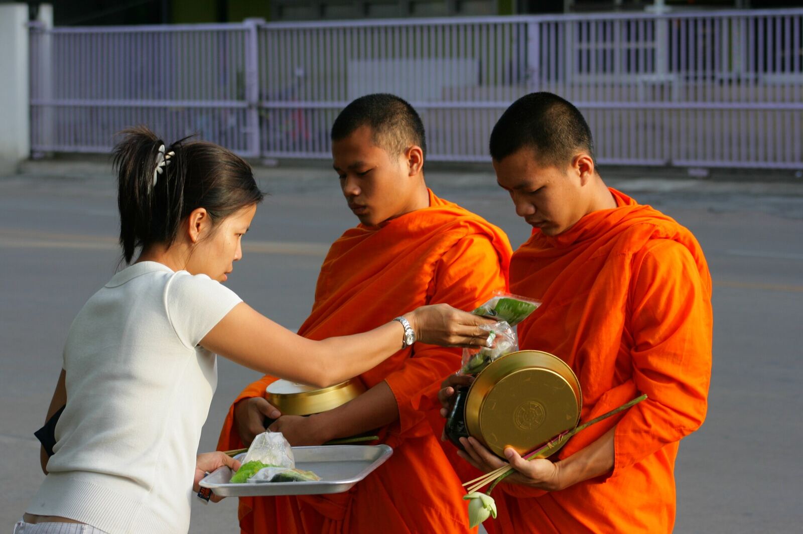 A woman in a white top places a packet of food into the alms bowl of one of two young monks in orange robes on the street.