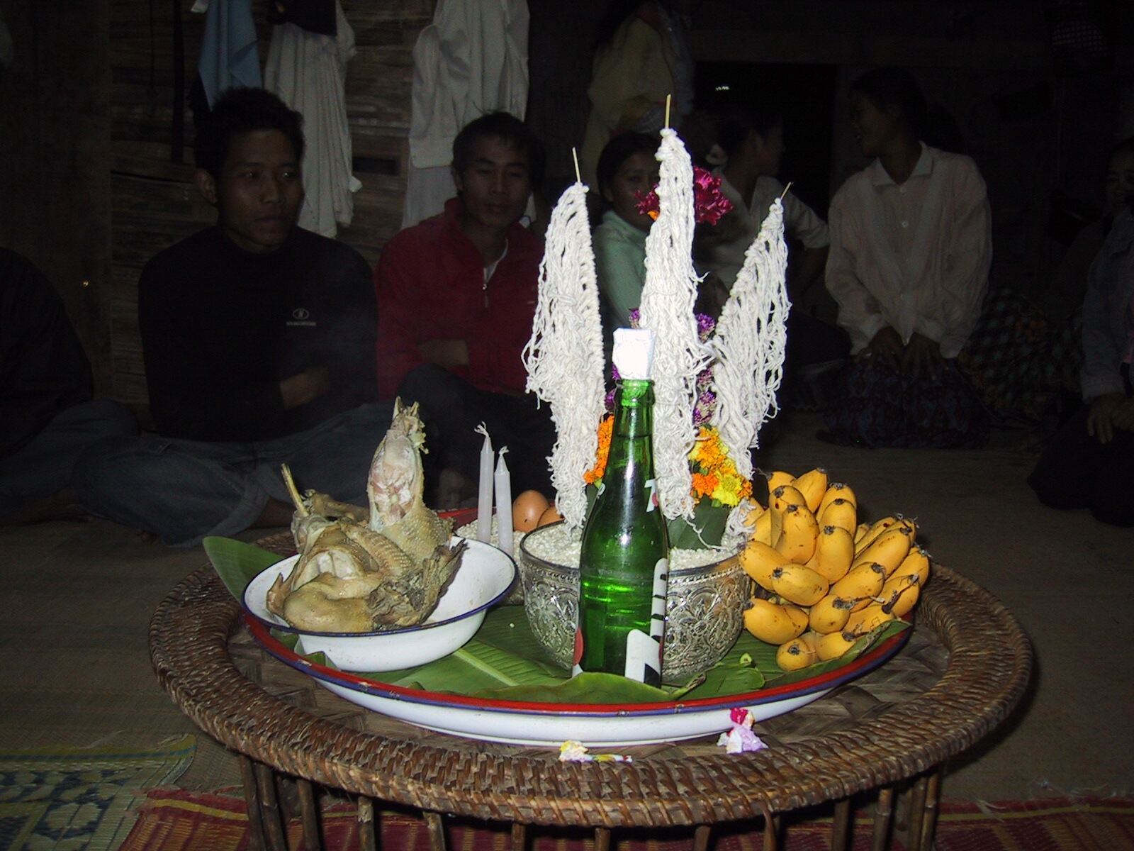 A round woven table holds a banana-leaf-lined tray with a boiled chicken, bananas, candles, a green bottle and tall white cotton-string cones, with villagers seated behind it.