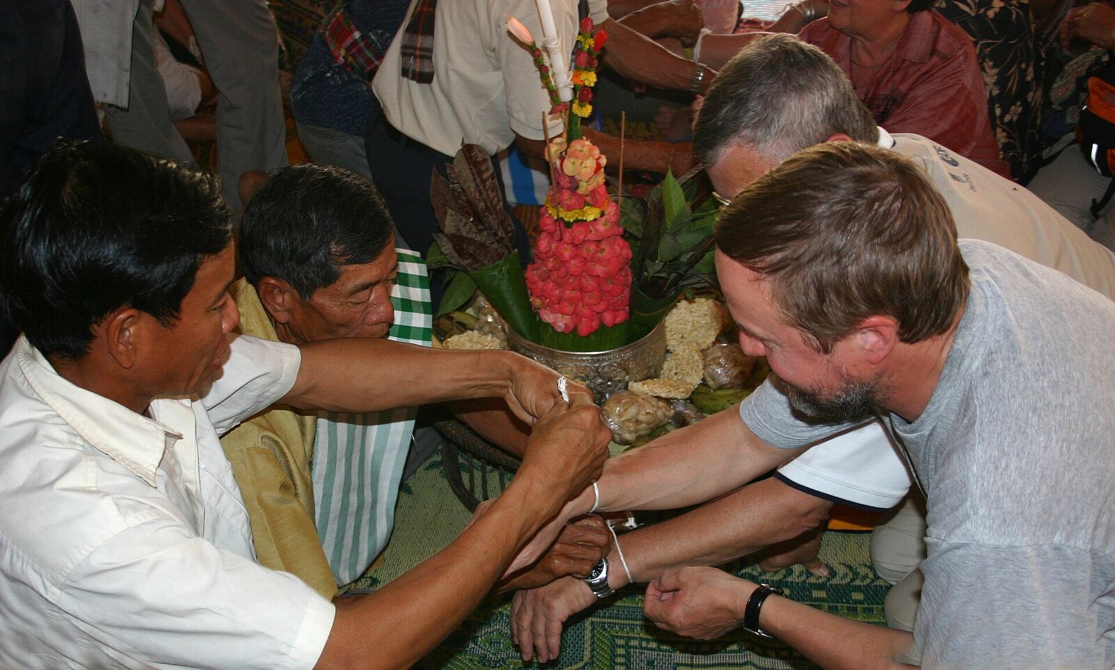 Lao villagers tie white cotton strings around the wrists of visitors who lean in over a small tray of flowers and offerings.