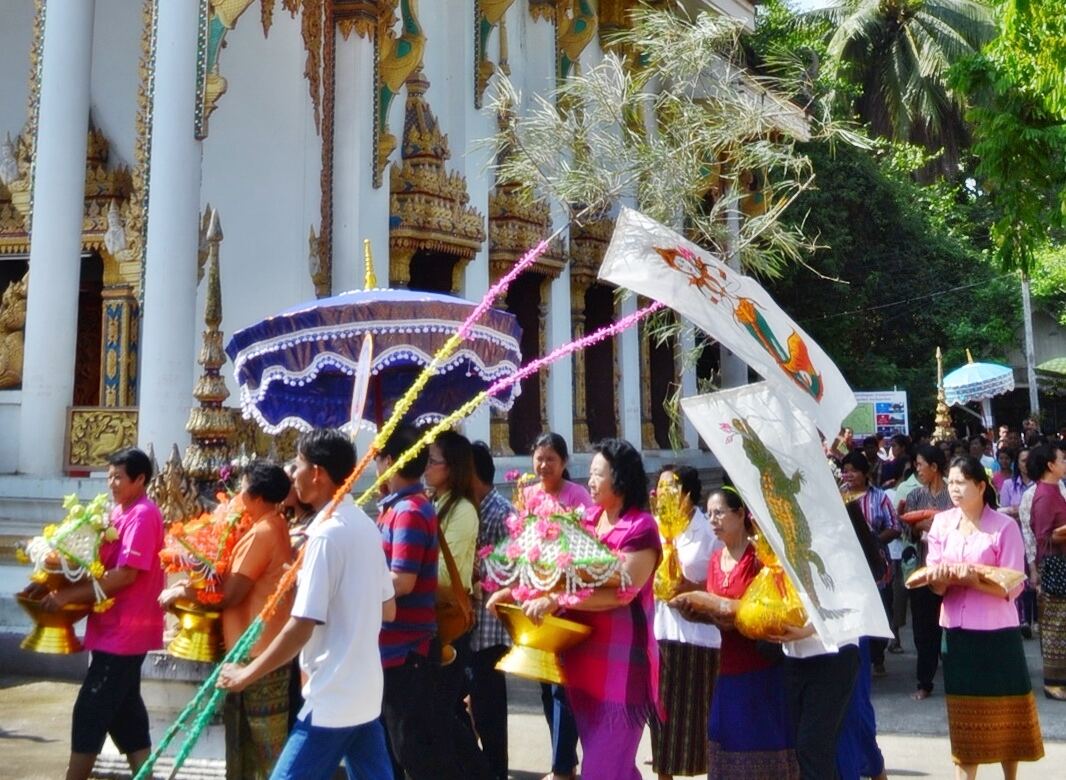 Villagers carry flower-decked offering stands and long painted banners in a Kathin procession in front of a temple hall, one under a blue parasol.