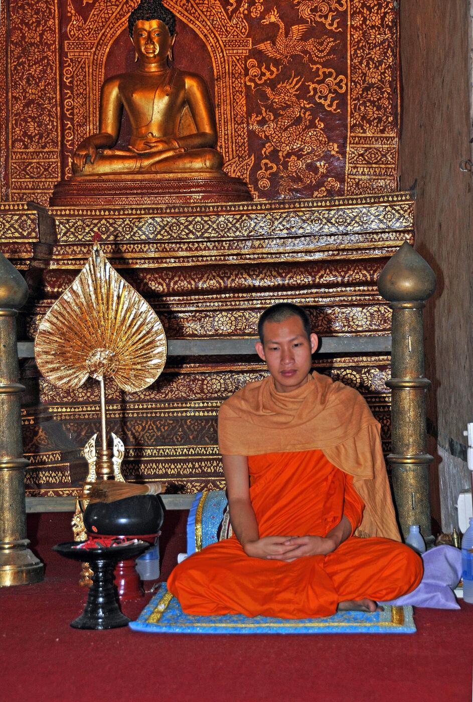A young monk sits cross-legged on a mat before a gilded Buddha in the Lai Kham viharn; the photographer's caption says he blessed visitors and tied a braided string on their wrists.