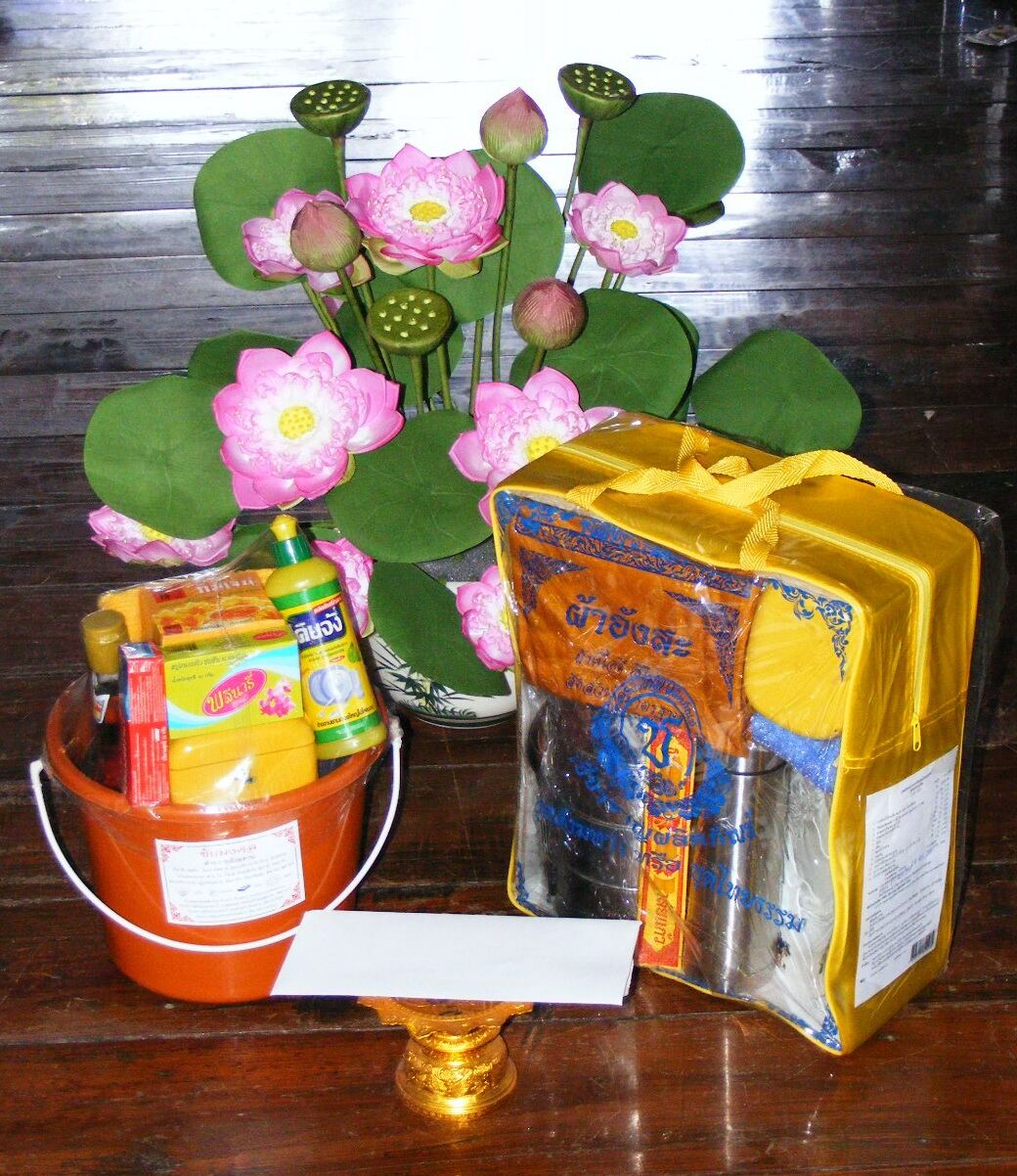 A sangkhathan offering set, an orange bucket of everyday goods and a packaged set of monk's requisites, stands beside pink lotus flowers on a polished table.