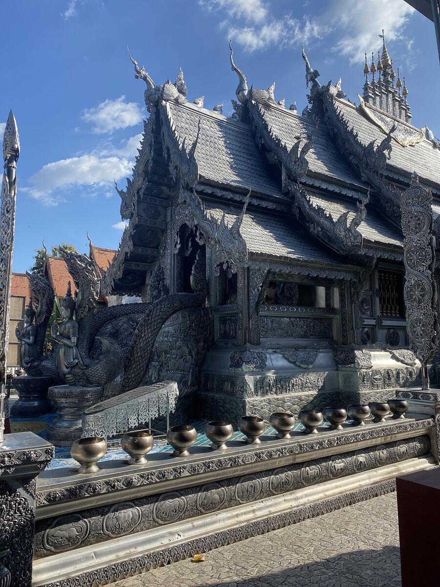 A row of dark silver bowls sits along a chased-silver platform in front of the temple's tiered silver roofs and naga stair rail.