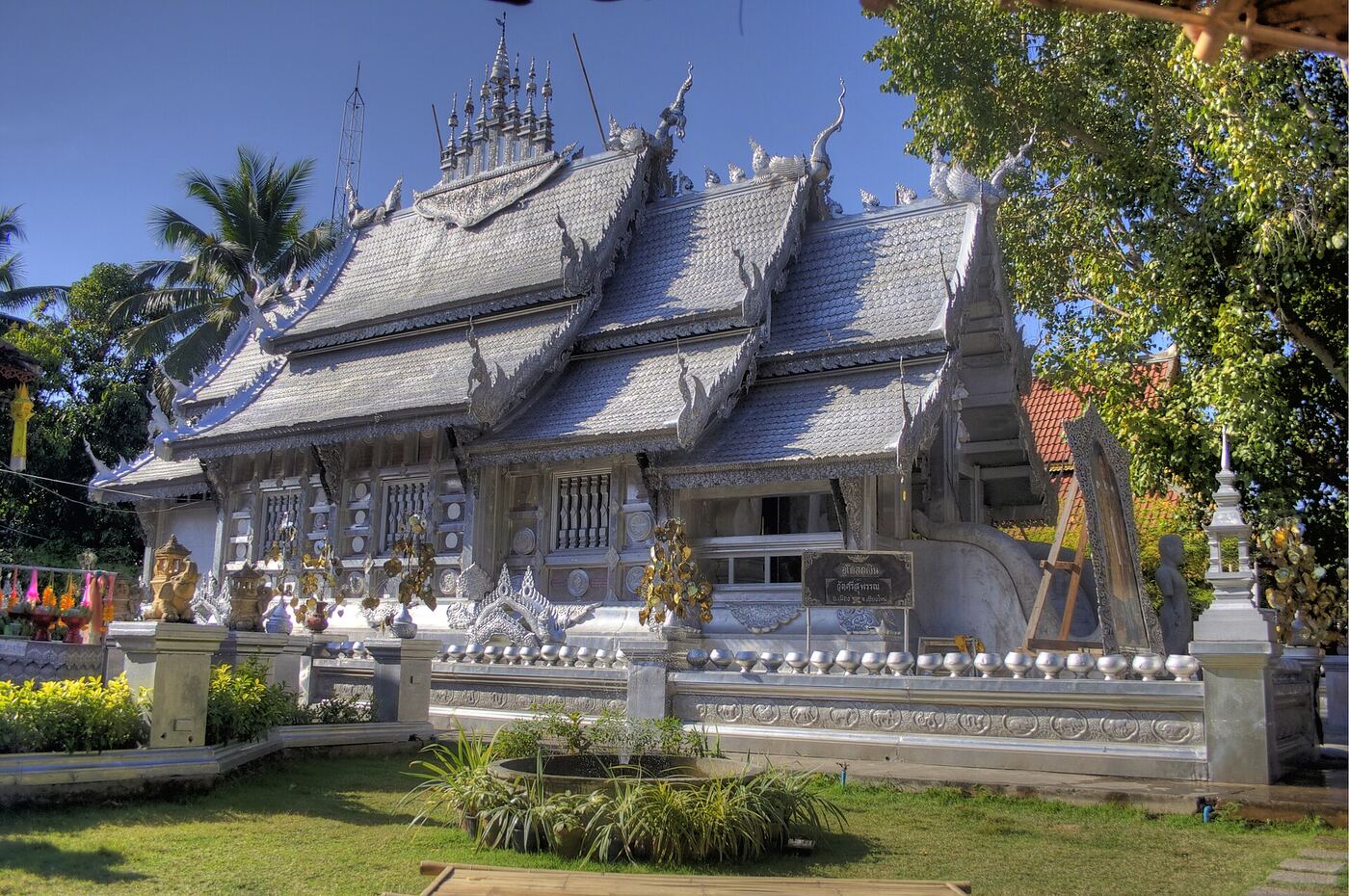 The silver-clad ubosot with tiered roofs stands behind a low wall, with a row of small silver bowls along its base.