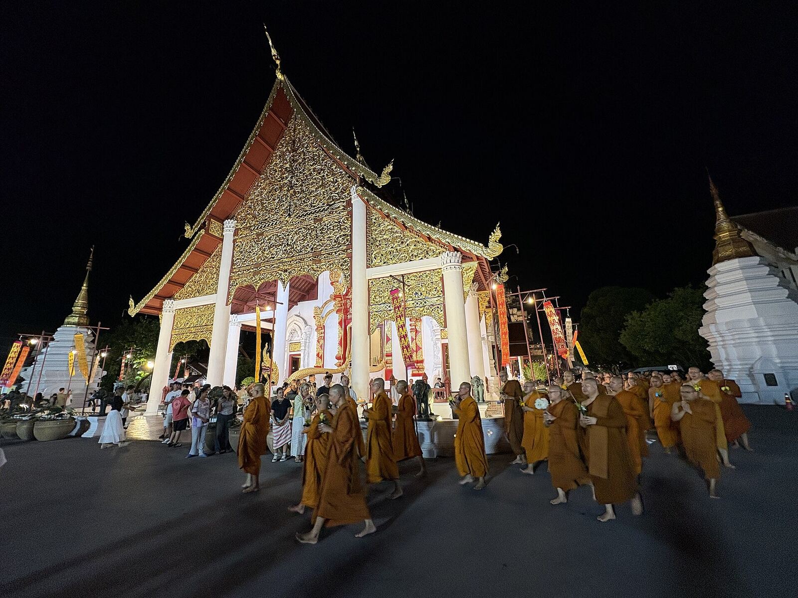 Monks in orange robes walk in procession past a gilded, white-pillared viharn lit against the night sky during the wian thian circumambulation.